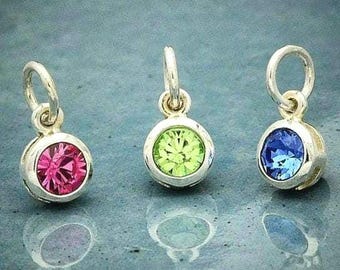May include: Three silver-toned metal charms, each featuring a circular bezel setting with a faceted gemstone. The left charm has a hot pink stone, the centre charm has a lime green stone, and the right charm has a bright blue stone.