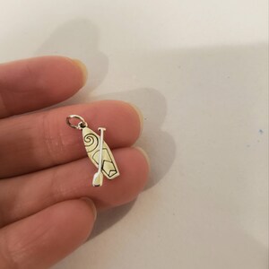 K&ouml;nnte beinhalten: Ein silberfarbener Anh&auml;nger mit einem Stand-Up-Paddleboard und Paddel. Das Paddleboard hat ein dekoratives Wirbeldesign. Der Anh&auml;nger ist klein und detailliert, mit einer Schlaufe oben zum Befestigen an einem Armband oder einer Halskette.