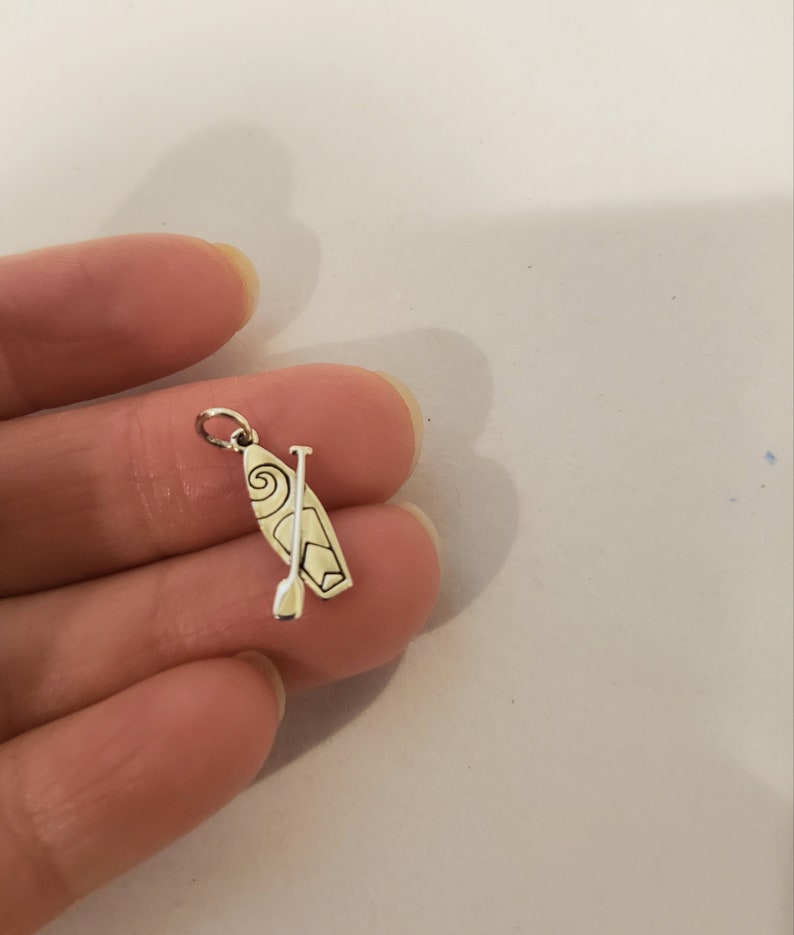 K&ouml;nnte beinhalten: Ein silberfarbener Anh&auml;nger mit einem Stand-Up-Paddleboard und Paddel. Das Paddleboard hat ein dekoratives Wirbeldesign. Der Anh&auml;nger ist klein und detailliert, mit einer Schlaufe oben zum Befestigen an einem Armband oder einer Halskette.