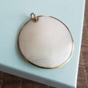 May include: A round, white mother of pearl pendant with a gold rim. The pendant has a loop at the top for attaching a chain.