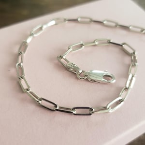 Puede incluir: Una pulsera de cadena plateada con diseño de eslabones ovalados. La pulsera tiene un cierre de mosquetón. La joya se muestra sobre una superficie rosa claro.