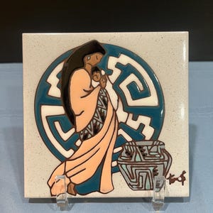 Peut inclure: Carreau de céramique carré avec un motif amérindien. L'œuvre représente une figure en robe pêche tenant un bébé, sur fond de motif circulaire bleu sarcelle et blanc. Un pot bleu sarcelle et gris est également représenté.
