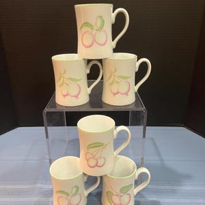 May include: A stack of six white ceramic mugs. Each mug features a pink cherry design with green leaves and stems. The rims and handles are outlined in green. The mugs are arranged on a clear acrylic stand and a blue tablecloth.