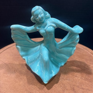 May include: A turquoise ceramic figurine of a woman in a flowing dress, dancing with her arms outstretched.