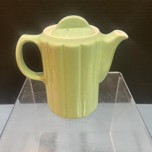 May include: A light green ceramic teapot featuring a vertical ribbed pattern along the cylindrical body, a matching lid with a small handle, a curved spout, and a side handle. The teapot has a smooth, glossy finish and a simple, functional design.