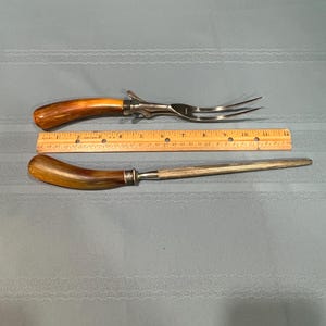 May include: A carving fork and a sharpening steel with wooden handles and stainless steel blades. The fork measures approximately 12 inches long. The sharpening steel is approximately 11 inches long. Both are displayed on a light blue tablecloth.