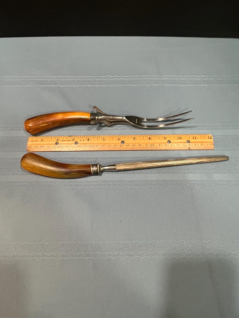 May include: A carving fork and a sharpening steel with wooden handles and stainless steel blades. The fork measures approximately 12 inches long. The sharpening steel is approximately 11 inches long. Both are displayed on a light blue tablecloth.