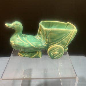May include: A ceramic duck planter in a vibrant teal color. The duck is pulling a small cart with a square opening. The cart has a wheel on each side. The planter is a decorative item.