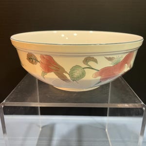 May include: A cream-colored ceramic bowl with a floral design. The bowl features red and pink flowers with green leaves and a light blue rim. The bowl is sitting on a clear acrylic stand.