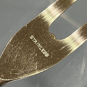 May include: Close-up of a stainless steel kitchen utensil. The word "STAINLESS" is etched into the metal. The utensil has a pointed end and a curved design with a hole.