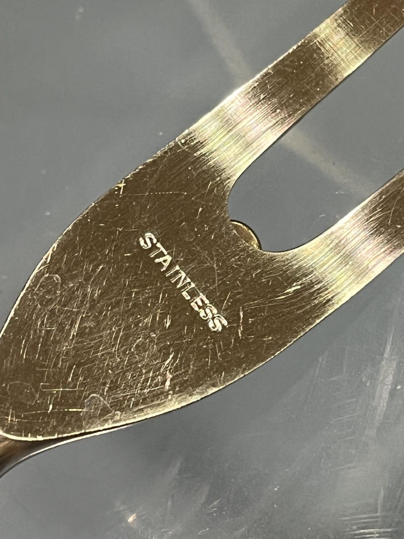 May include: Close-up of a stainless steel kitchen utensil. The word "STAINLESS" is etched into the metal. The utensil has a pointed end and a curved design with a hole.