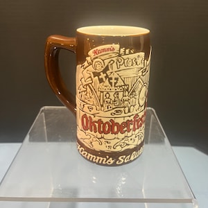 May include: A brown ceramic beer stein with a handle, featuring the Hamm's logo and the word "Oktoberfest" in red script. The stein has a cream-colored interior and a detailed illustration of a festive scene.
