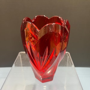 May include: A vibrant red glass vase with a unique, scalloped rim and a faceted, tulip-like shape. The vase has a geometric design with pointed, vertical cuts that catch the light, creating a sparkling effect. The vase is displayed on a clear acrylic surface.