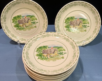 Può includere: Un set di piatti in ceramica color crema con un bordo di foglie verdi. Ogni piatto presenta un'immagine centrale di una scena di cottage con un tetto giallo e fogliame verde. I piatti sono impilati e disposti su una superficie blu.