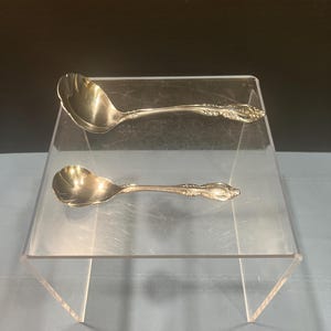 May include: Two silver-toned metal spoons with scalloped edges and ornate floral detailing on the handles. The larger spoon is positioned above the smaller spoon, both featuring a polished, reflective finish and a curved, shell-like bowl shape.