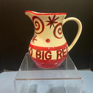 May include: A white ceramic pitcher with a red rim and handle. The pitcher has a red and black swirl design and the words "So Big Red" in black lettering on a red band.