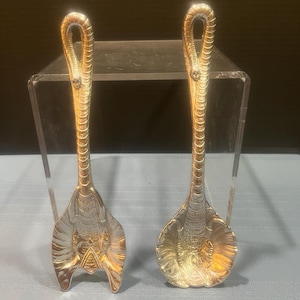 May include: Two silver-toned metal spoons featuring long, textured handles that curve into loops at the top. The bowls of the spoons are embossed with detailed elephant head designs, with the trunks extending upward to form the stems of the spoons.