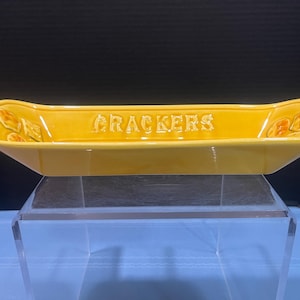 May include: Yellow ceramic cracker dish with the word "CRACKERS" in raised lettering. The dish has a rectangular shape with rounded ends and decorative leaf accents. A vintage serving piece.