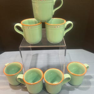 May include: A set of six green ceramic mugs with orange rims and handles. Each mug has a decorative band with a small design near the top. The mugs are stacked on a clear display stand.