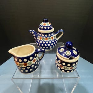 May include: A three-piece ceramic tea set with a blue and white polka dot pattern, featuring a teapot, creamer, and sugar bowl. The set has a floral design with red and green accents. The teapot has a lid and spout.
