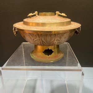 May include: A decorative brass bowl with a lid, featuring intricate floral and geometric patterns. The bowl has a round base and handles on the sides. The color is a mix of gold and bronze.