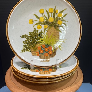 May include: Set of four ceramic dinner plates with a floral design featuring yellow tulips, green leaves, and orange flowers in woven baskets. The plates have a speckled white background with a brown rim.