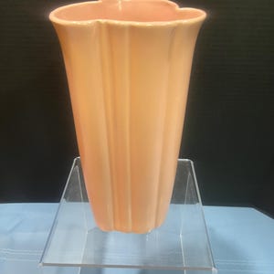 May include: A peach-colored, fluted vase with a scalloped rim. The vase is displayed on a clear acrylic stand against a dark background. The vase has vertical ridges and a flared shape, suitable for floral arrangements.