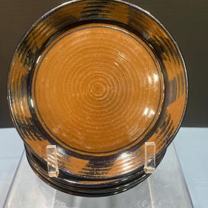 May include: A stack of round ceramic plates featuring a smooth, matte, terracotta-colored center with concentric circular ridges. The wide, glossy rim is finished in a deep, dark navy blue glaze with visible brushstroke patterns along the edge.
