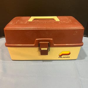 May include: A brown and beige plastic Plano tackle box with a latch and a handle. The box has the Plano logo on the front.