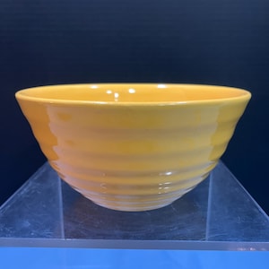 May include: A bright yellow ceramic bowl with a glossy finish. The bowl has a rounded shape with horizontal ridged detailing around the exterior. The bowl is sitting on a clear, angled surface against a dark blue background.
