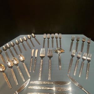 May include: A collection of vintage silverware, including spoons, forks, knives, and butter knives, arranged on a light blue surface. The silverware has ornate, engraved handles and a shiny, metallic finish, suggesting a formal dining set.
