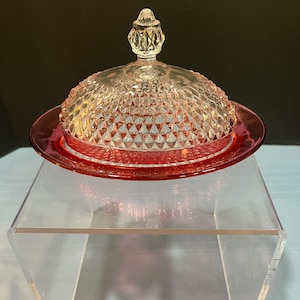 May include: A vintage glass butter dish with a ruby red base and a clear, diamond-patterned dome lid. The lid has a decorative finial. The dish sits on a clear acrylic stand.