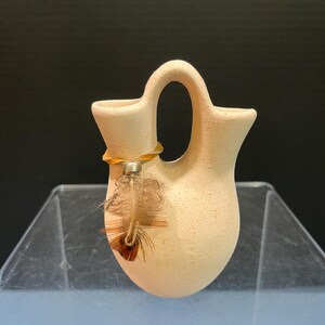 May include: A small, cream-colored ceramic vessel with a textured, matte finish. The piece features a unique double-spout design connected by a central arched handle. A thin tan leather cord is tied around one spout, adorned with a silver bead and a brown feather.