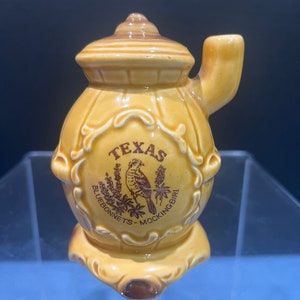 May include: A small, yellow ceramic teapot shaped like a wood-burning stove with a brown handle and a brown lid. The teapot has a brown design with the words "Texas Bluebonnets - Mockingbird" and a brown image of a mockingbird on a branch with bluebonnets.