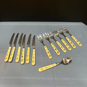 May include: A collection of flatware with cream-colored handles featuring a green cactus design. The set includes knives, forks, and a spoon. The utensils are arranged on a light blue tablecloth.