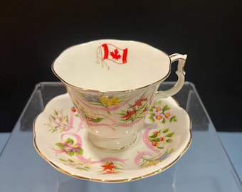 May include: A white teacup and saucer set with gold trim. The teacup features a small Canadian flag and floral designs. The saucer has a scalloped edge and a matching floral pattern with pink ribbons. A decorative tea set.