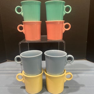 May include: Six ceramic mugs stacked in pairs. The top two mugs are turquoise, the middle two are pink, and the bottom two are light blue and yellow.