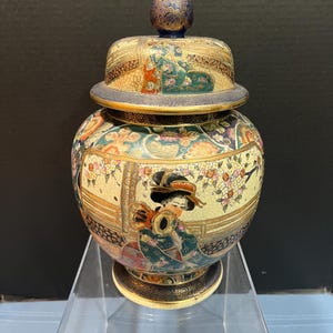 May include: A ceramic Japanese-style jar with a lid. The jar is decorated with gold and black floral patterns, and a scene of a woman in a kimono. The lid is decorated with a gold and black floral pattern.
