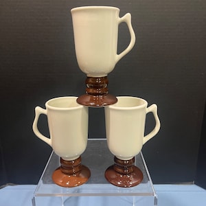 May include: Three white ceramic mugs with brown pedestal bases. The mugs are stacked on top of each other.