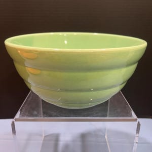 May include: A light green ceramic bowl with a rounded shape and a slightly flared rim. The bowl has three horizontal ridges around the exterior. The bowl is sitting on a clear acrylic stand.