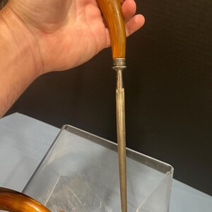 May include: A vintage knife sharpener with a curved, amber-colored handle and a silver-colored metal bolster. The sharpening steel is long and slender. The handle is held in a hand.