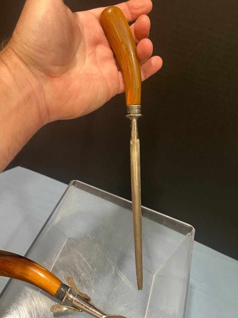 May include: A vintage knife sharpener with a curved, amber-colored handle and a silver-colored metal bolster. The sharpening steel is long and slender. The handle is held in a hand.