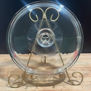 May include: A clear glass lid with a decorative pattern on the underside. The lid is on a gold metal stand with a round top and three legs.