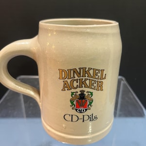 May include: A beige ceramic beer stein with a handle. The stein features the words "DINKEL ACKER" in gold lettering, along with a crest and the words "CD-Pils" in black. The stein has a slightly textured surface.