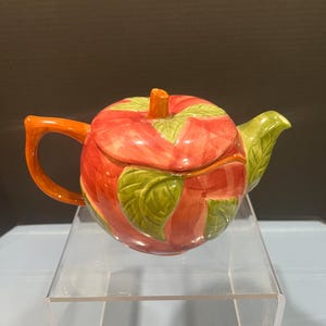 May include: A ceramic teapot shaped like a red apple with green leaves and a brown stem. The handle and spout are orange and green. The teapot is displayed on a clear acrylic stand.
