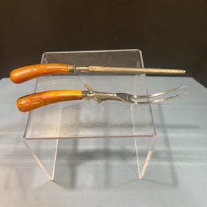 May include: A carving set with a sharpening steel and a carving fork. The handles are a warm brown color, and the metal is silver. The carving fork has two tines. The set is displayed on a clear acrylic stand.