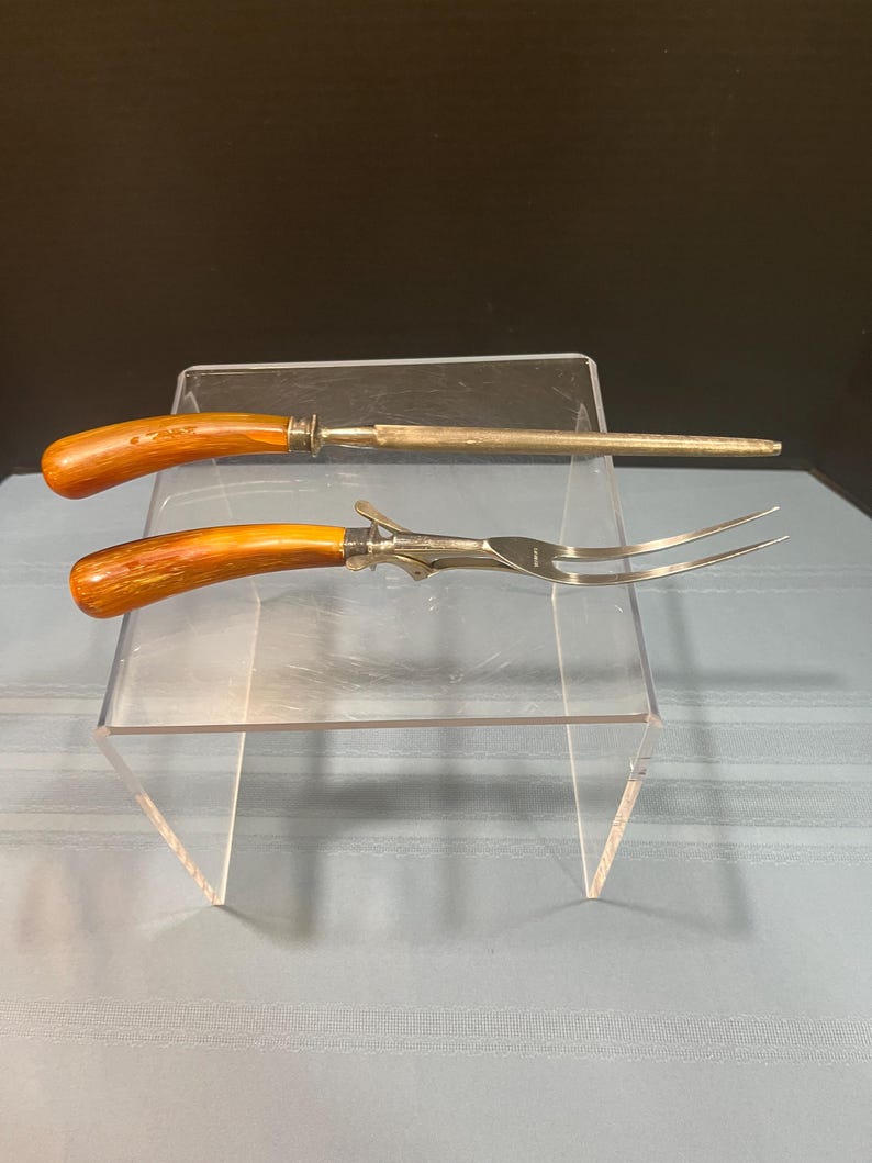 May include: A carving set with a sharpening steel and a carving fork. The handles are a warm brown color, and the metal is silver. The carving fork has two tines. The set is displayed on a clear acrylic stand.