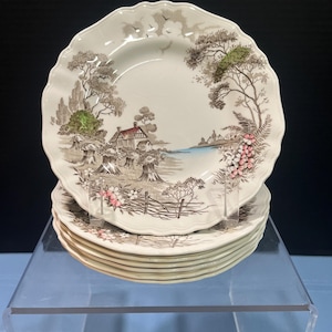 May include: A stack of cream-colored ceramic plates with scalloped edges. Each plate features a detailed brown transferware landscape scene depicting a cottage, trees, wheat sheaves, and a fence, with accents of green and pink floral details.