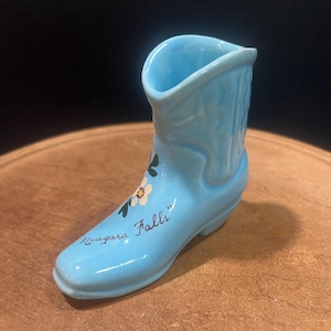 May include: A light blue ceramic boot with white flowers painted on the side. The boot has "Niagara Falls" written on the bottom.