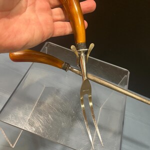 May include: A carving fork and serving fork set with brown handles and silver-colored metal. The carving fork has two tines, and the serving fork has a long, thin handle. The set is displayed on a clear acrylic surface.